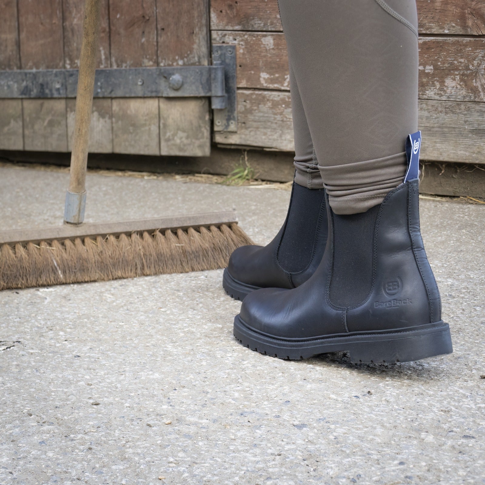 Person wearing black leather boots Bareback Boots, standing on a concrete surface with a wooden door in the background.
