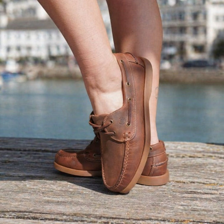 Brown loafers worn by a person standing on a wooden dock with a harbor background