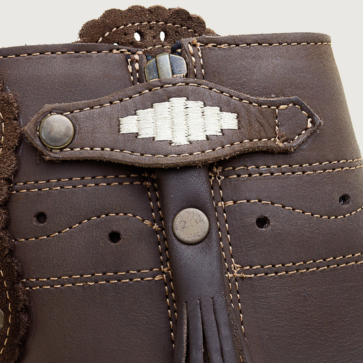 Close-up of a dark brown leather boot or mule featuring a decorative strap closure with a white embroidered geometric pattern, metal rivets, and contrast stitching.